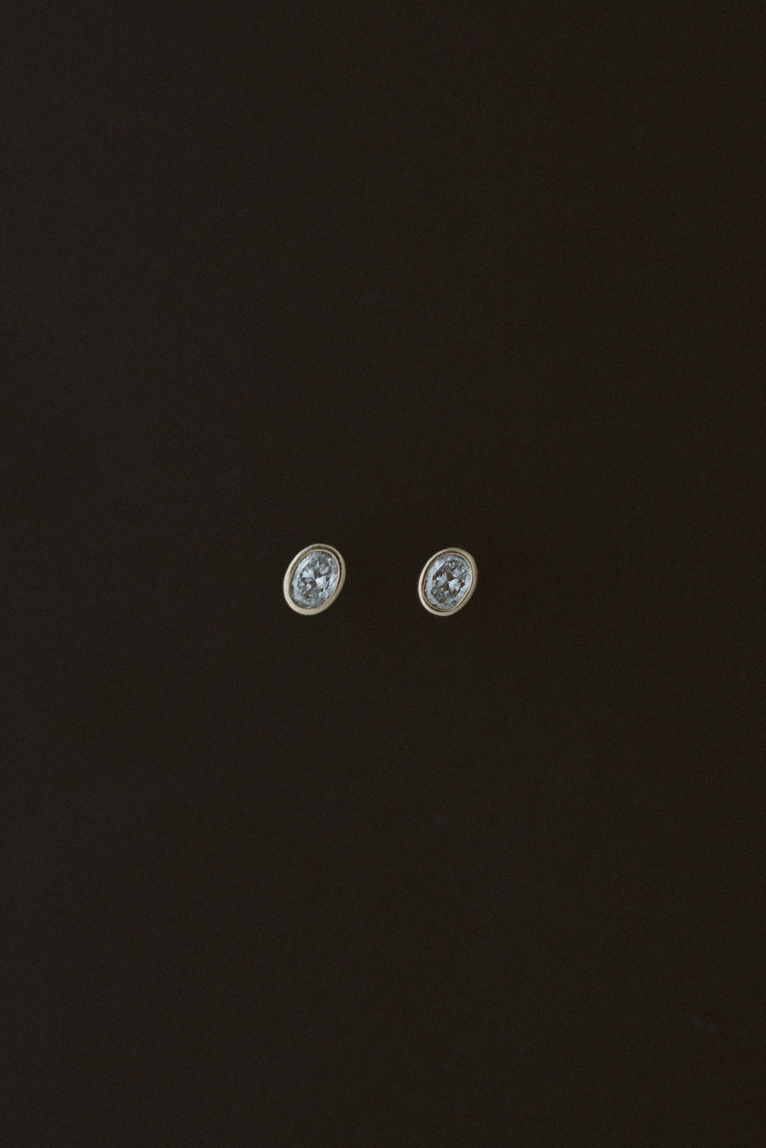 Oval Damsel Studs, bezel set oval diamond earrings in 14kt yellow gold on a dark background