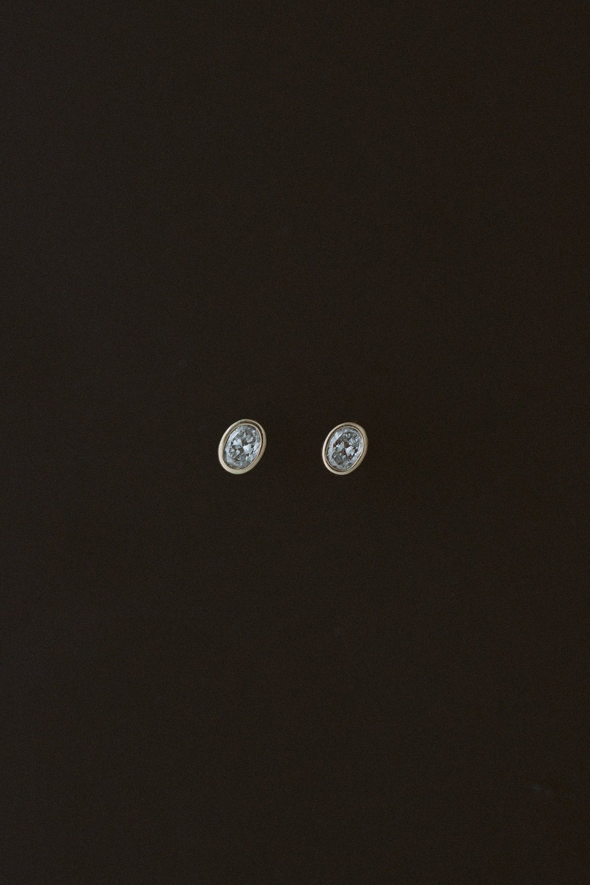 Oval Damsel Studs, bezel set oval diamond earrings in 14kt yellow gold on a dark background