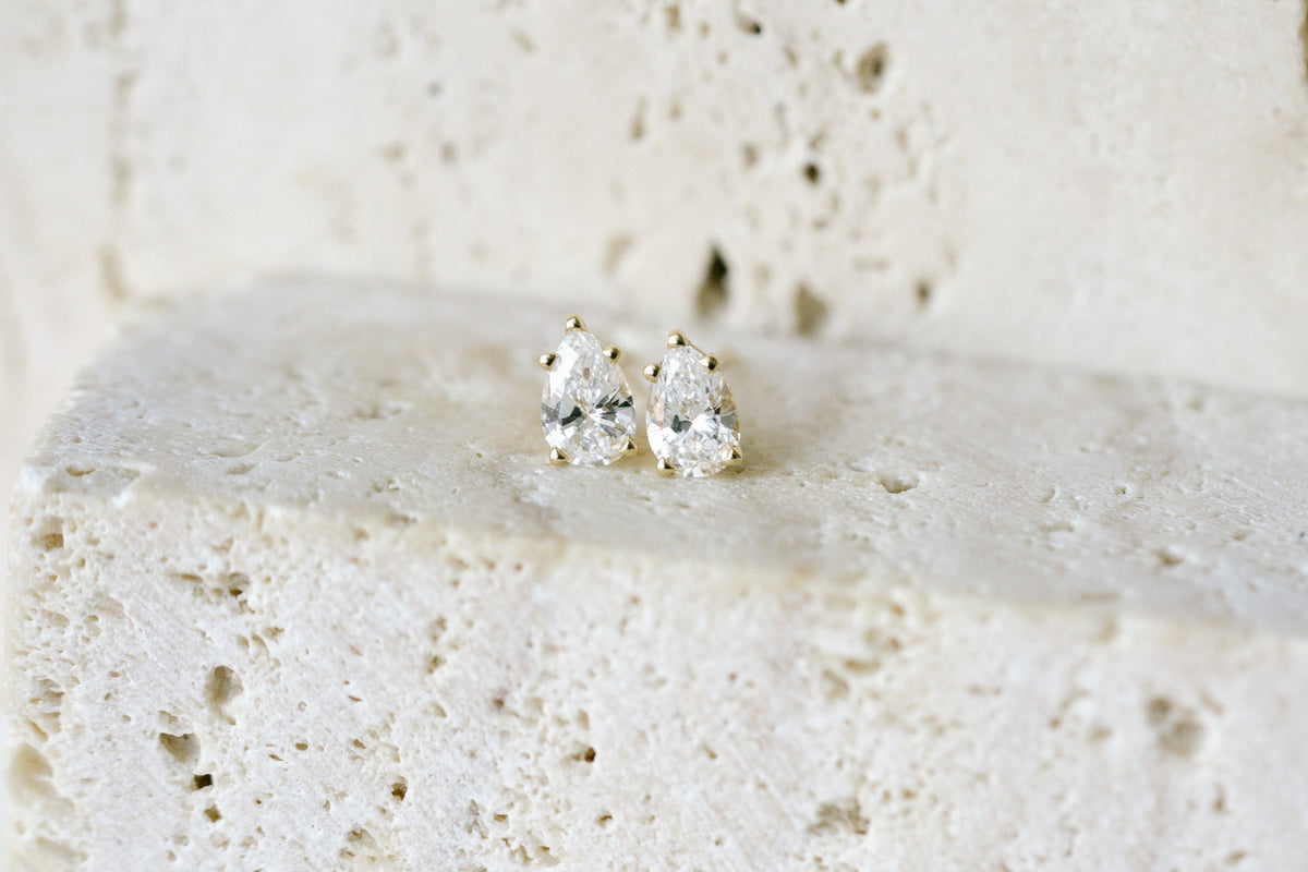BB Prong Pear Dew Drops, pear diamond studs in a 14kt yellow gold prong basket on a travertine ledge