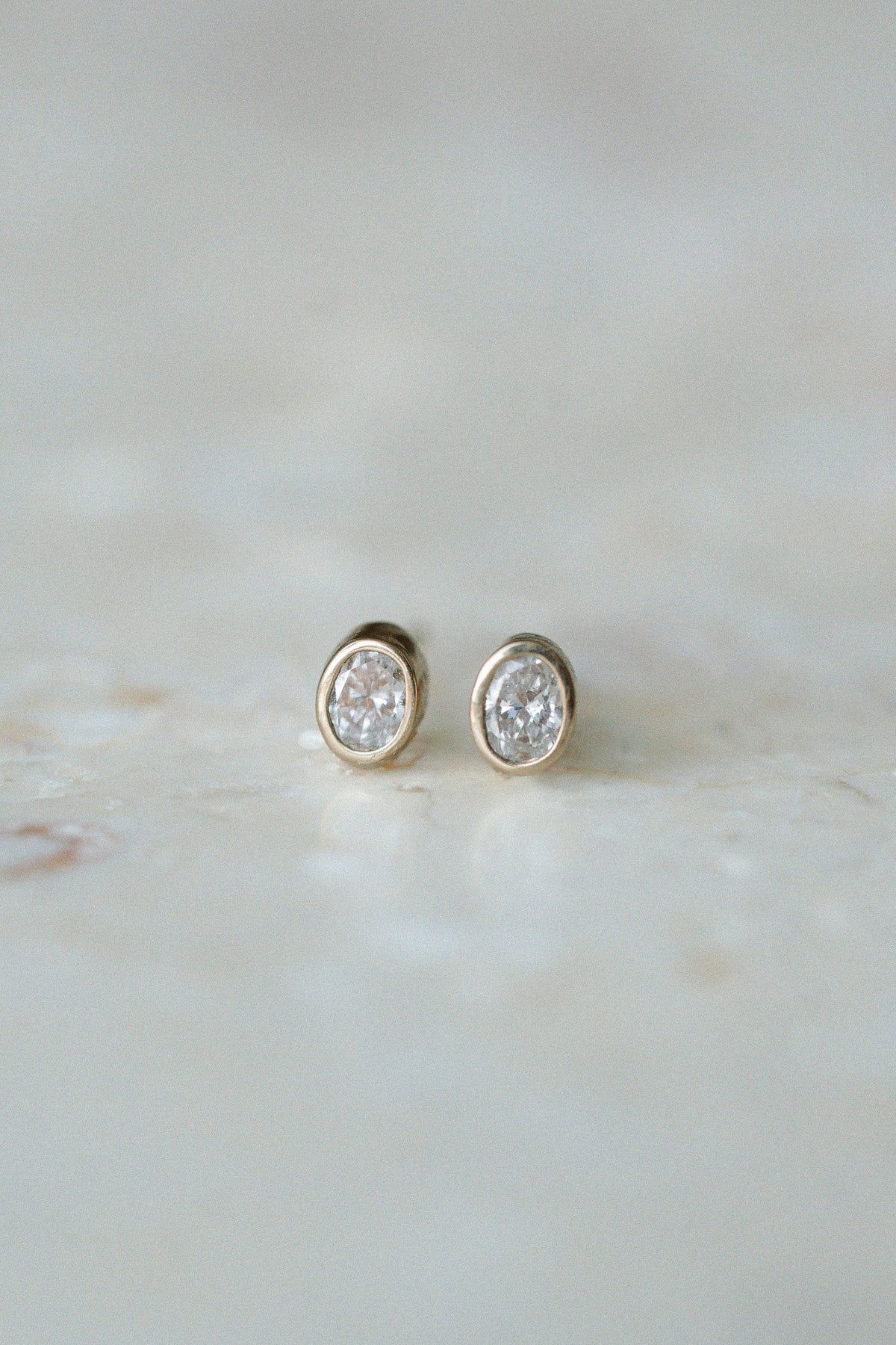 Oval Damsel Studs, bezel set oval diamond earrings in 14kt yellow gold on stone, close up